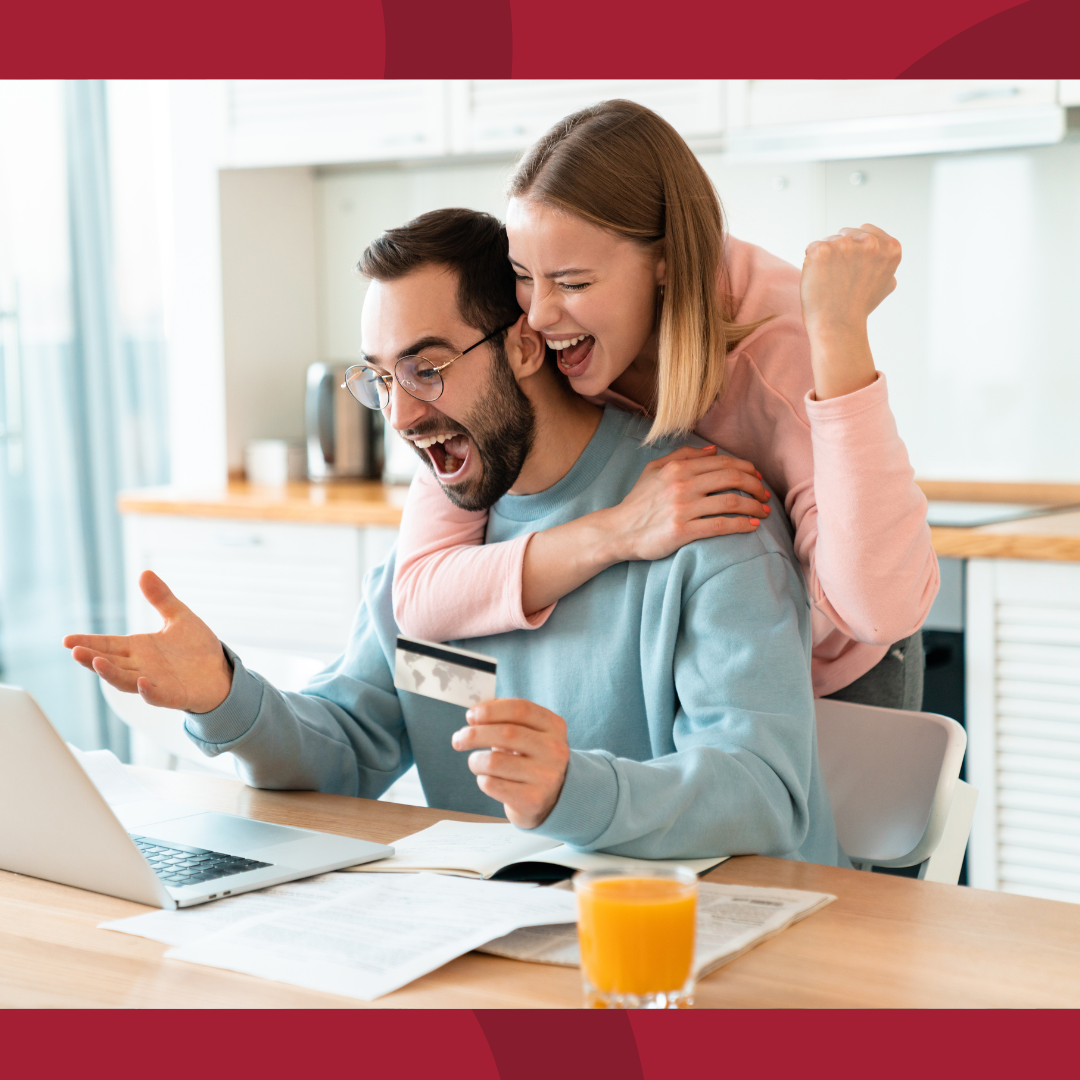 A man is sitting at the kitchen table in front of a laptop with woman leaning around him.  Both are excited about what is on the laptop.